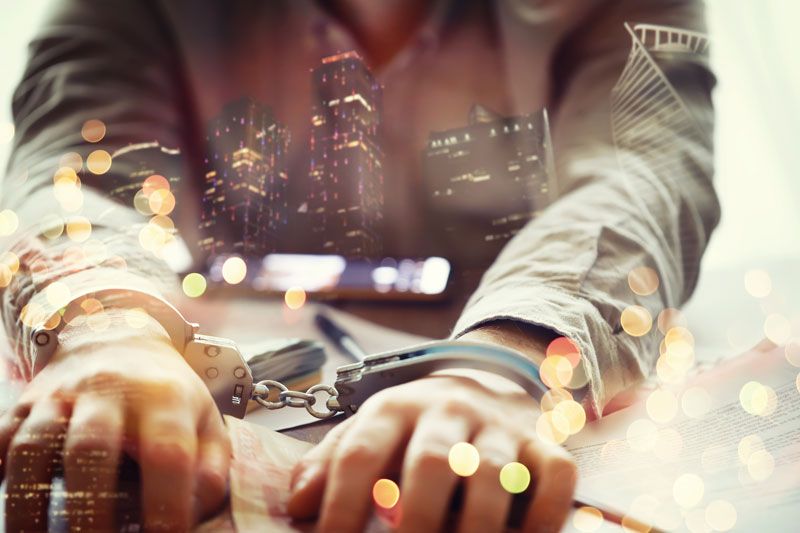 Hands in handcuffs resting on papers on a desk, with a night city skyline and bokeh lights blended over the photo