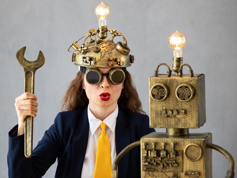 A businesswoman in a brass helmet with a light bulb and dark goggles holds a wrench next to a cardboard robot